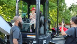 Child in the cab of excavator as caregiver takes photo and public works employee looks on