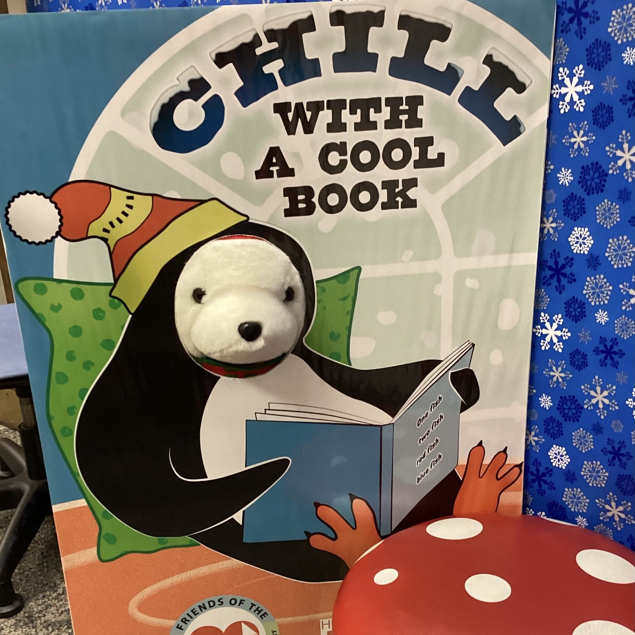 A stuffed polar bear using a selfie cutout of a penguin reading a book beside a stuffed mushroom stool
